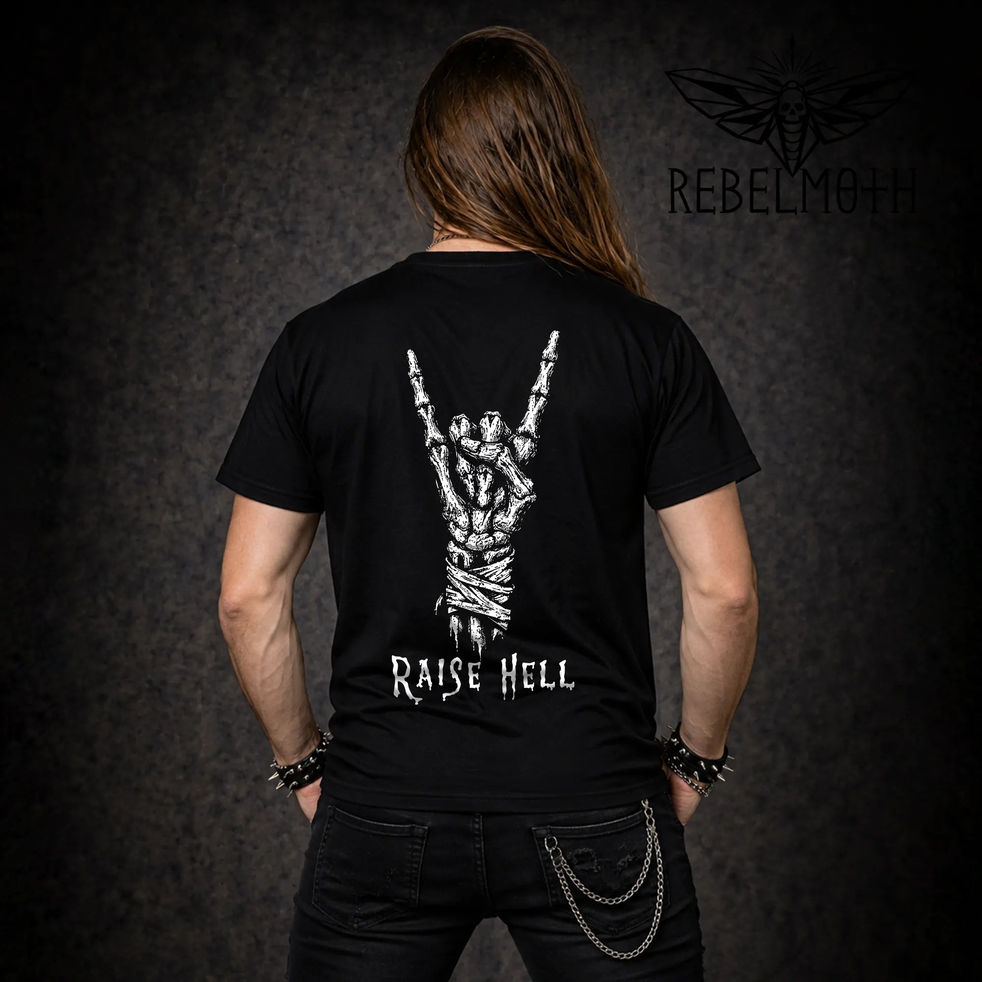 Back view of black t-shirt with skeleton hand throwing devil horns and Raise Hell text, worn by long-haired model with spiked bracelets and chain wallet